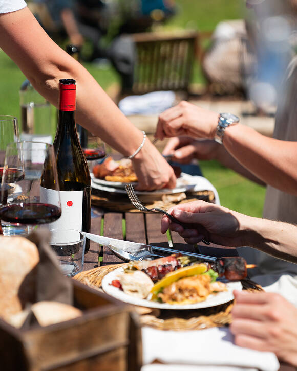 Mani di commensali che condividono un'abbondante bistecca con osso affettata, servita con vino rosso durante un evento di catering BBQ a Trento.