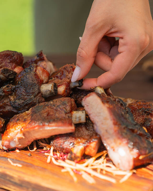 Mano di un ospite che prende una succulenta costina di carne da un tagliere in legno durante un catering BBQ a Trento.