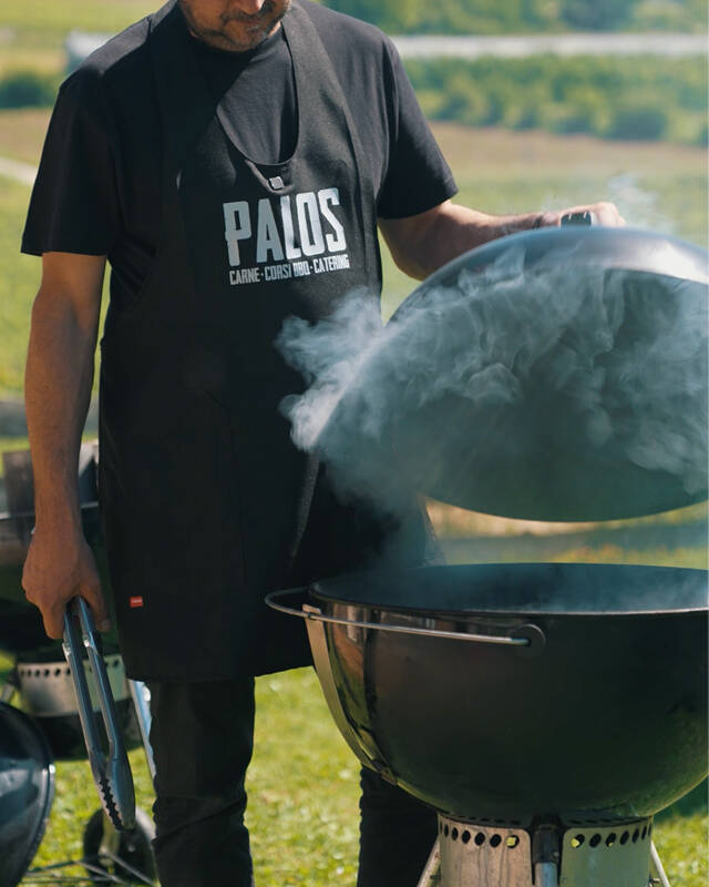 Un esperto con grembiule Palos solleva il coperchio di un affumicatore liberando una nuvola di fumo durante un corso di barbeque all'aperto a Trento.