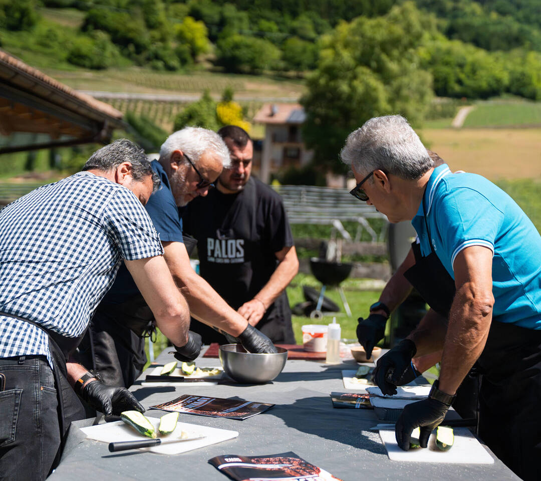 Quattro uomini con i guanti preparano del cibo su un tavolo all’aperto, su cui sono disposti taglieri, libri e una ciotola, circondati dal verde e dalle colline.