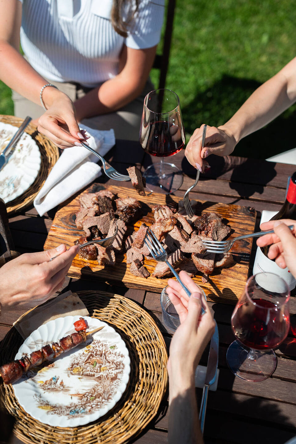 Persone che condividono carne BBQ su un tagliere durante un catering all'aperto a Trento, accompagnato da vino rosso.