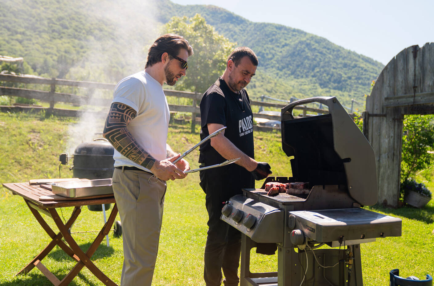 Due uomini preparano la carne su un grande barbecue a gas all'aperto durante un corso pratico di BBQ organizzato da Palos Barbeque a Trento, con le montagne sullo sfondo.