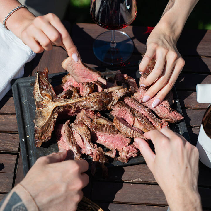 Mani di commensali che condividono un'abbondante bistecca con osso affettata, servita con vino rosso durante un evento di catering BBQ a Trento.